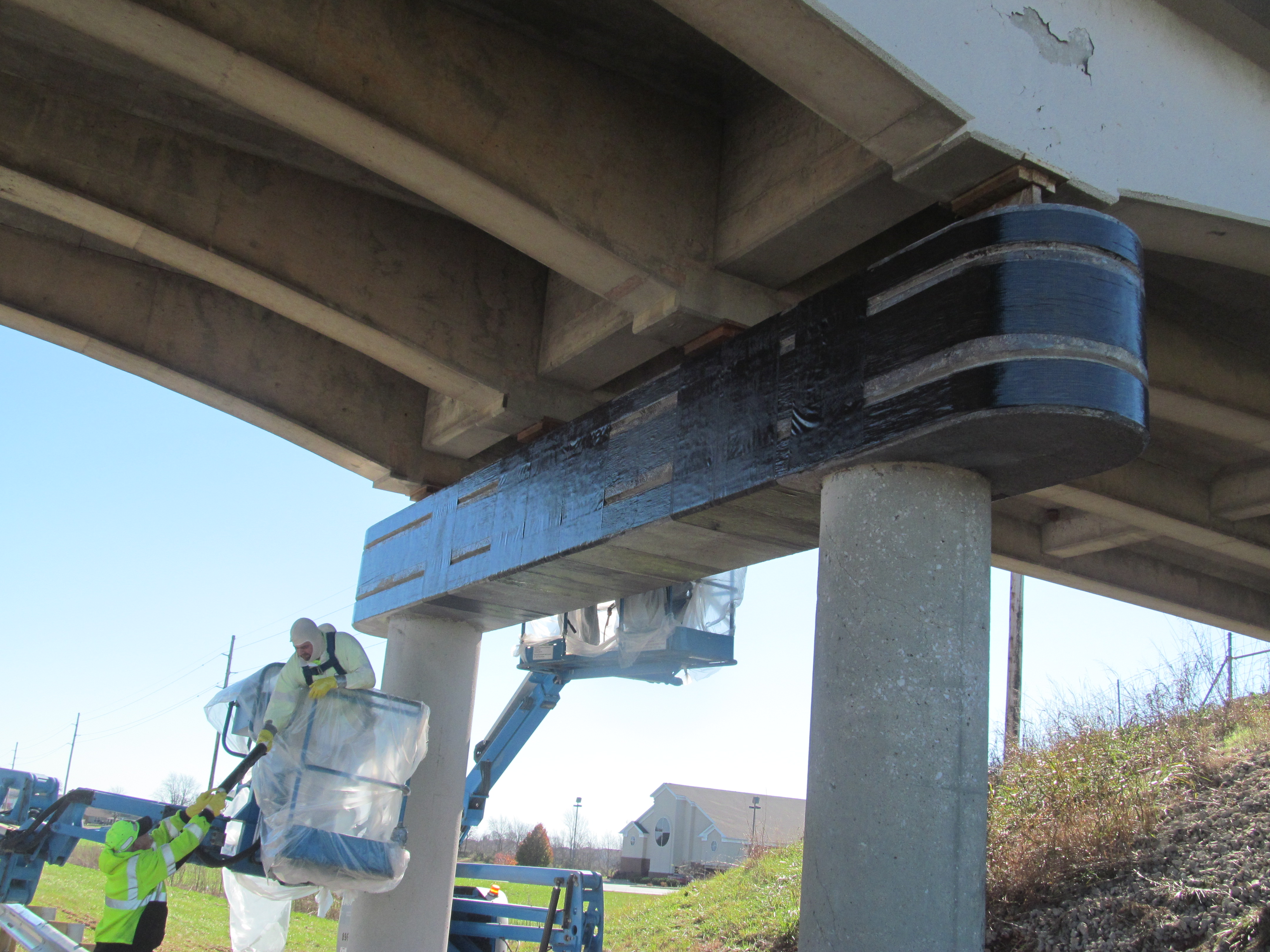 Photo of bridge pier cap being wrapped with carbon fabric. Two sets of manlifts are being used. The manlift in front has two construction workers in white coveralls. 