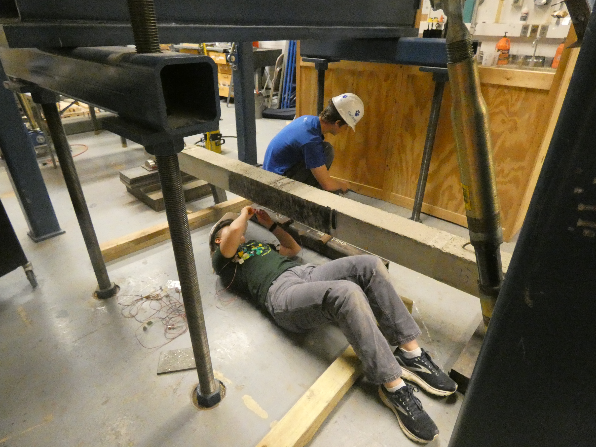 Photo of two students with white hard hats working on a concrete beam on supports in the structures lab. One student is laying on her back attaching strain gauge wire, while the other student is running the wired under a wooden shield. 