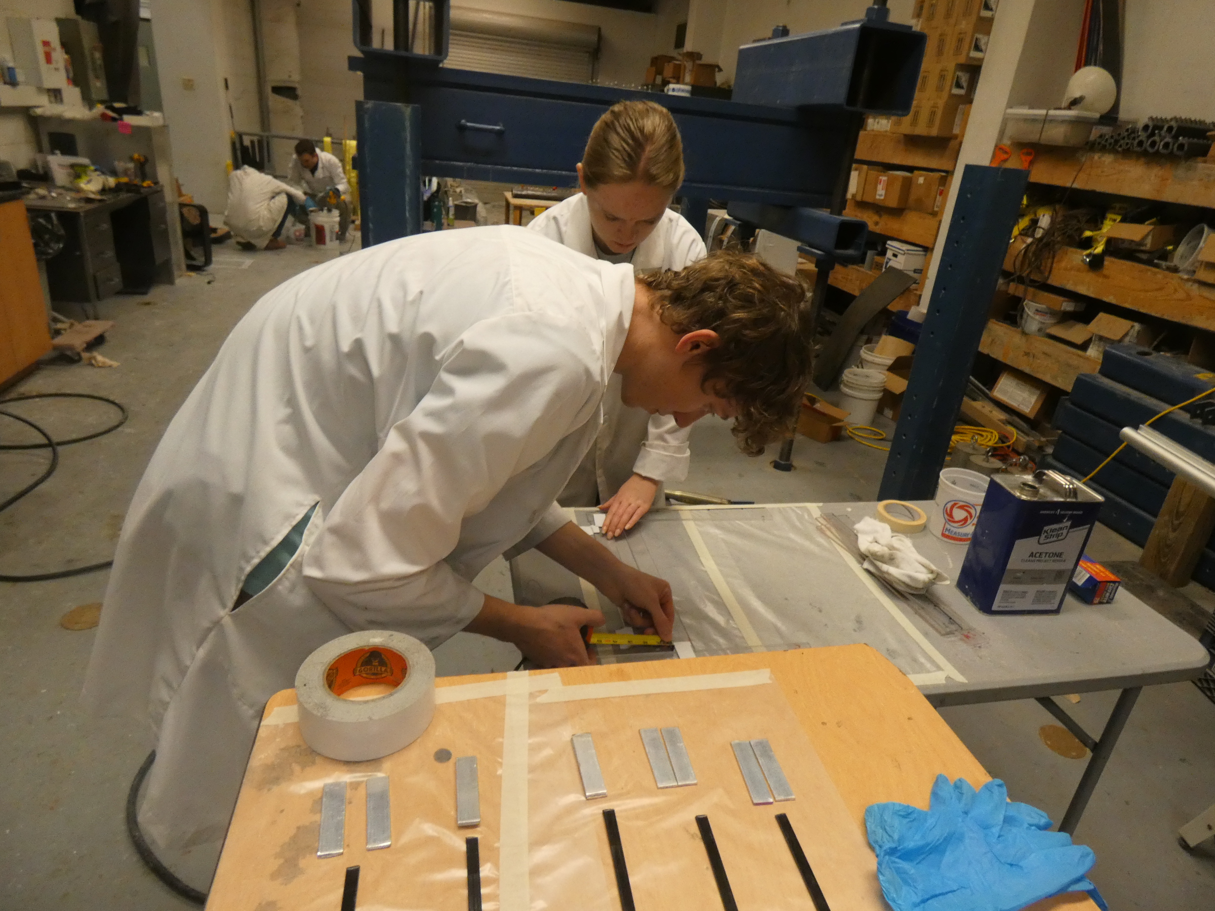 Photo of two students in the foreground wearing white lab coats preparing FRP specimens for testing. There are two other students in white lab coats in the background mixing epoxy. 