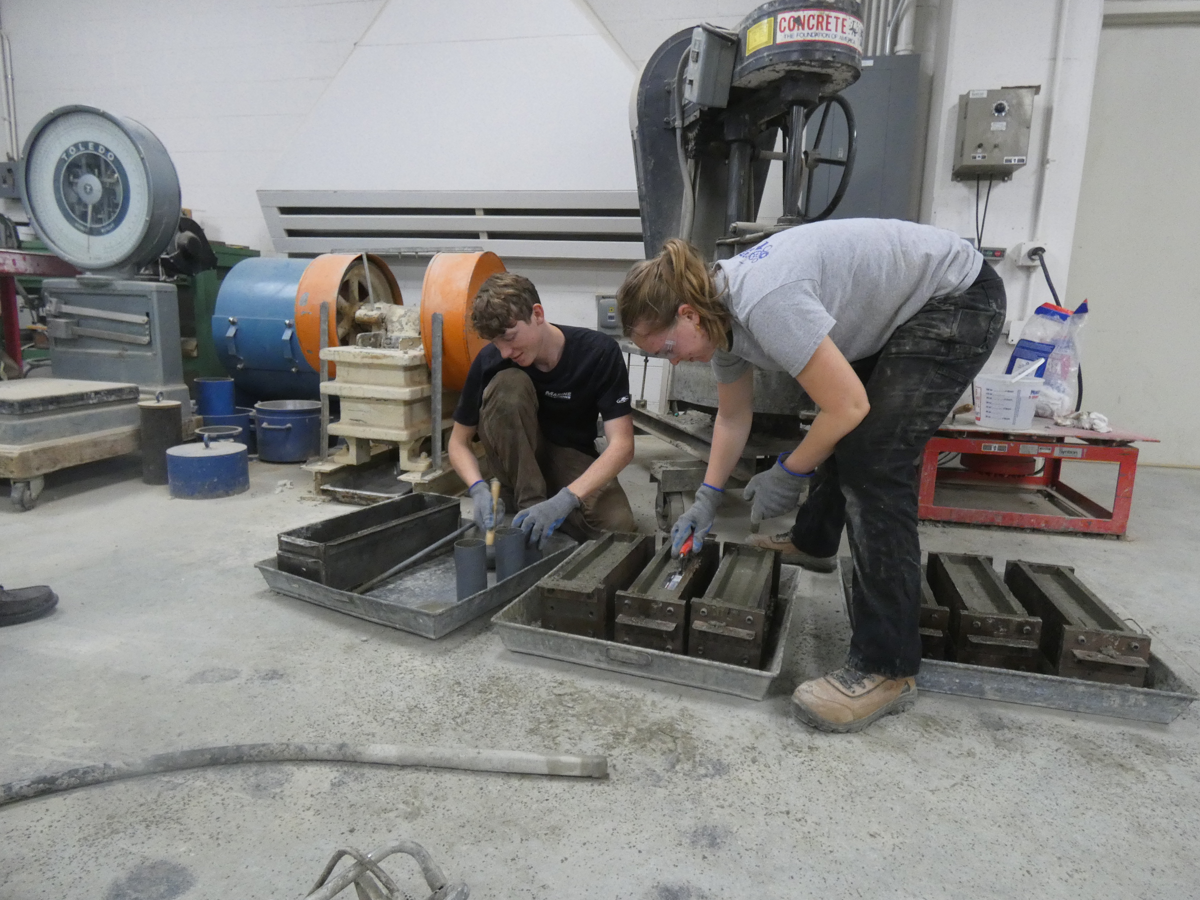 Photo of two student placing concrete in molds. One is preparing small beams, while the other is making cylinders. 