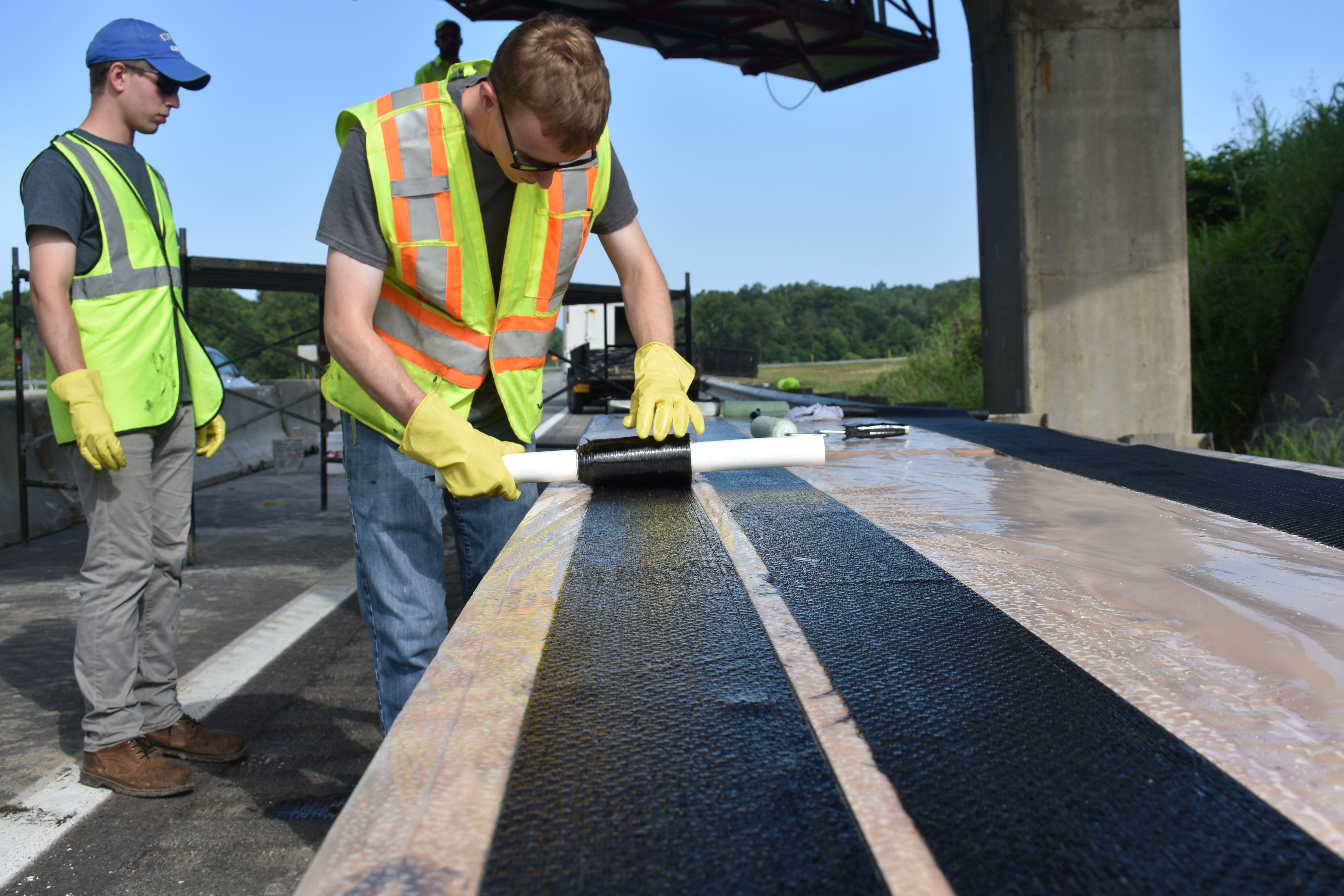 Photo of student rolling a saturated carbon fabric sheet while another student watches on underneath a bridge. 