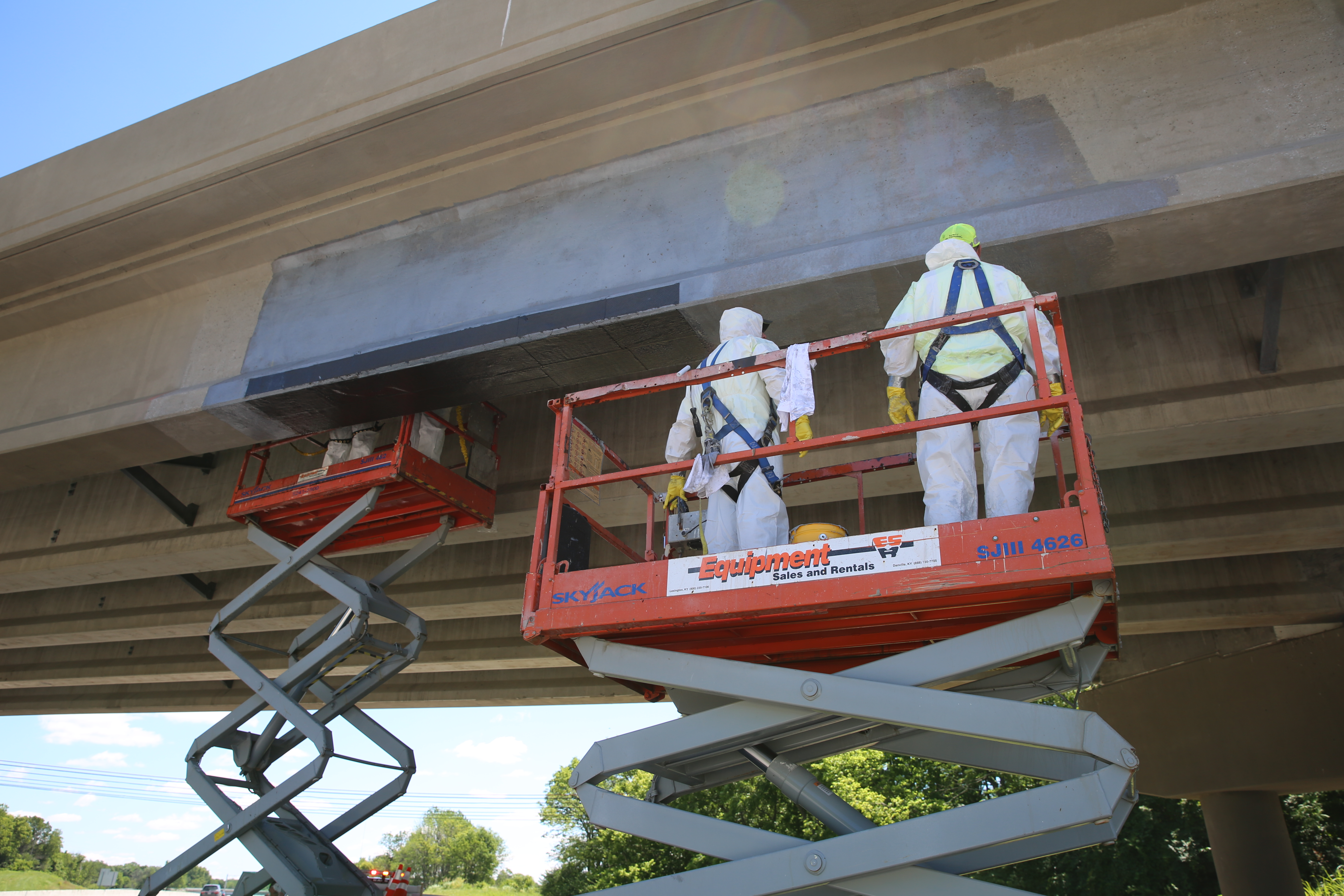 Photo of construction workers applying carbon fabric on to the bottom flange of a prestressed I-beam. There are two scissor lifts with two workers in white coveralls in each lift. 