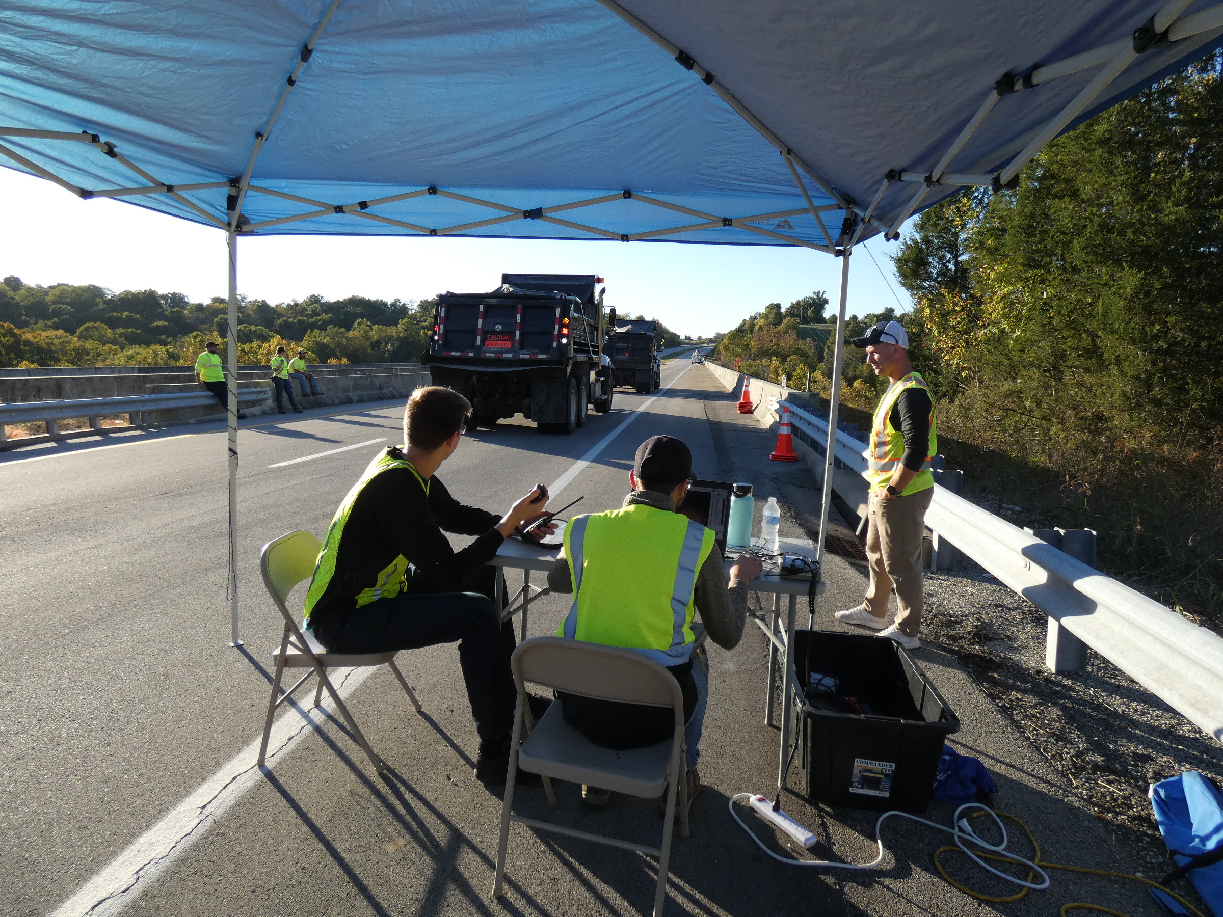 Photo of two students seated with a laptop on a table in front of them by the side of the road next to a bridge with a KYTC bridge crew member standing next to them. The bridge is being loaded using two tandem rear axle dump trucks. 