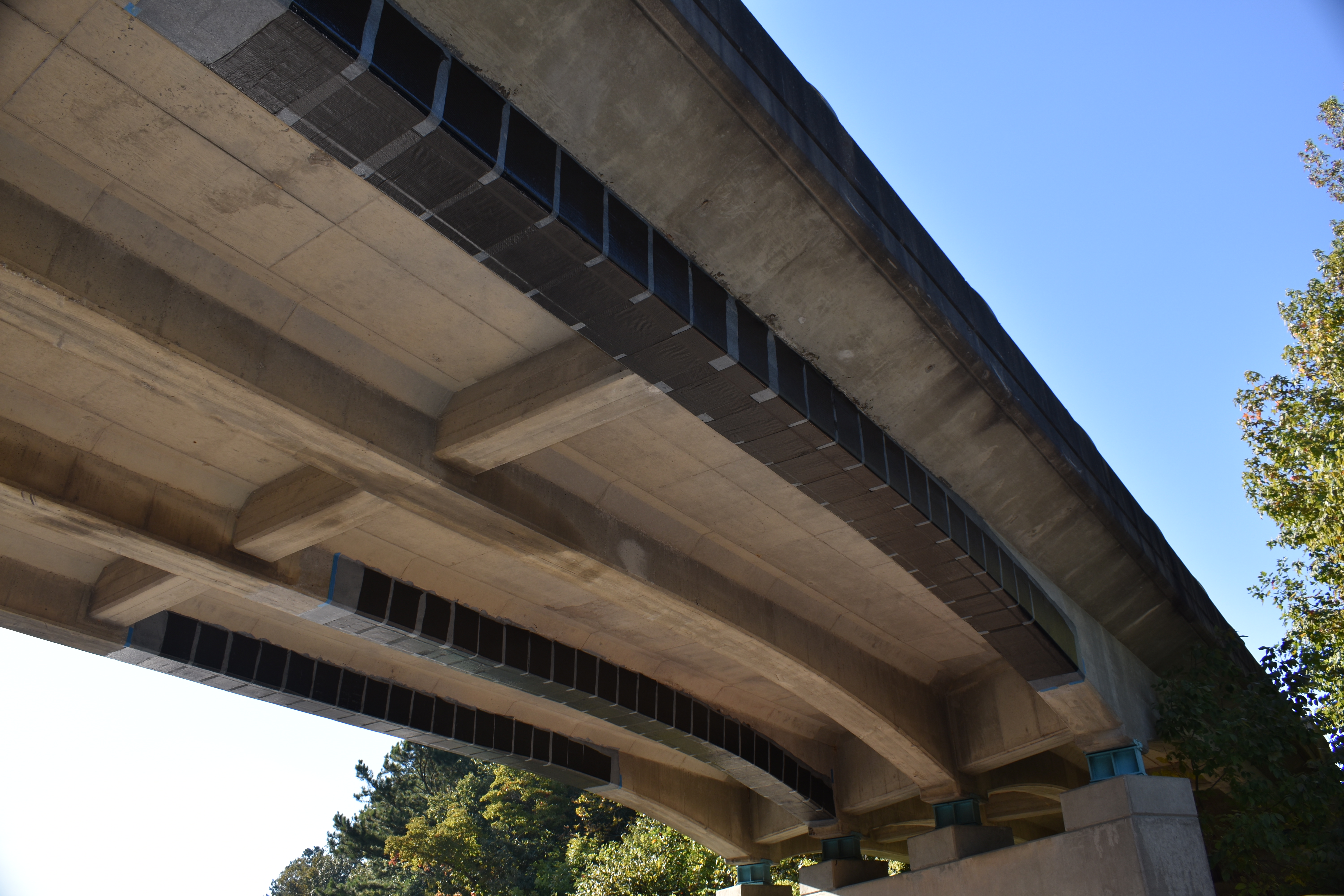 Photo of underside of a span in a reinforced concrete bridge with four beams. Three of the beams are wrapped with carbon fabric. 