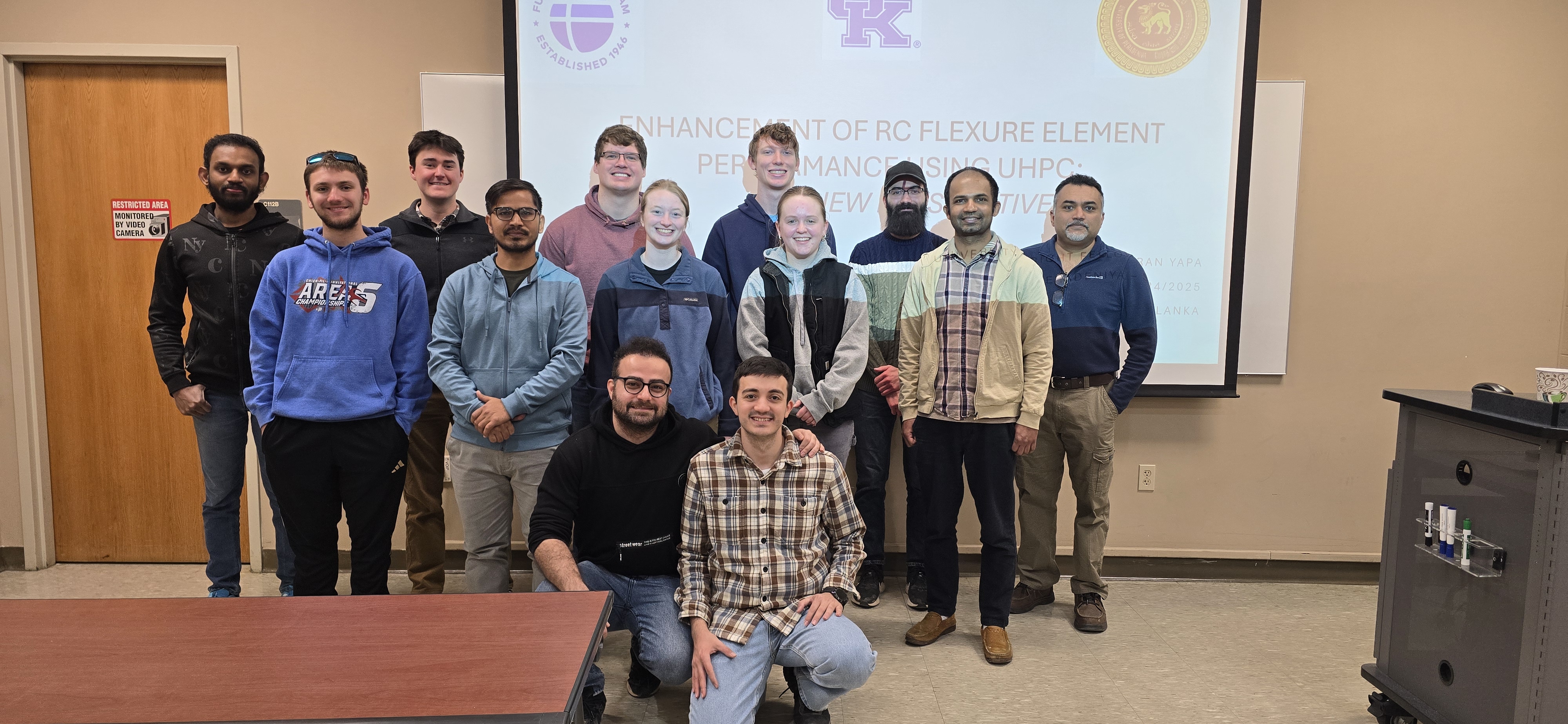 Photo of Dr. Abheetha Peiris, nine students and a visiting professor standing in front of a screen in two rows. Two other students are kneeling in front.