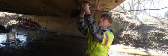 Photo of student applying a strain gauge on the bottom of beam in a reinforced concrete bridge.   